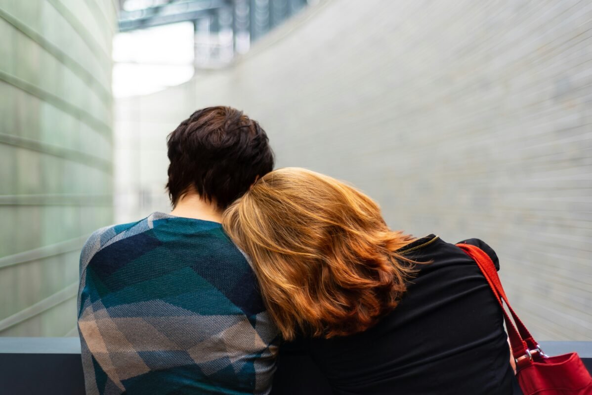 Two people seen from behind, one resting their head on the other's shoulder, walking through a quiet corridor
