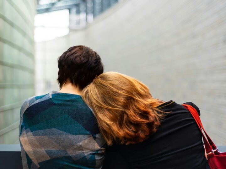 Two people seen from behind, one resting their head on the other's shoulder, walking through a quiet corridor